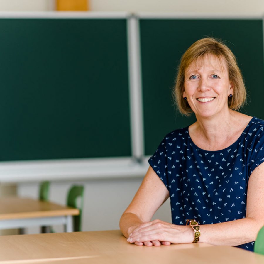 Birgit Kilian, sitzend an einem Schultisch mit einer Schultafel im Hintergrund